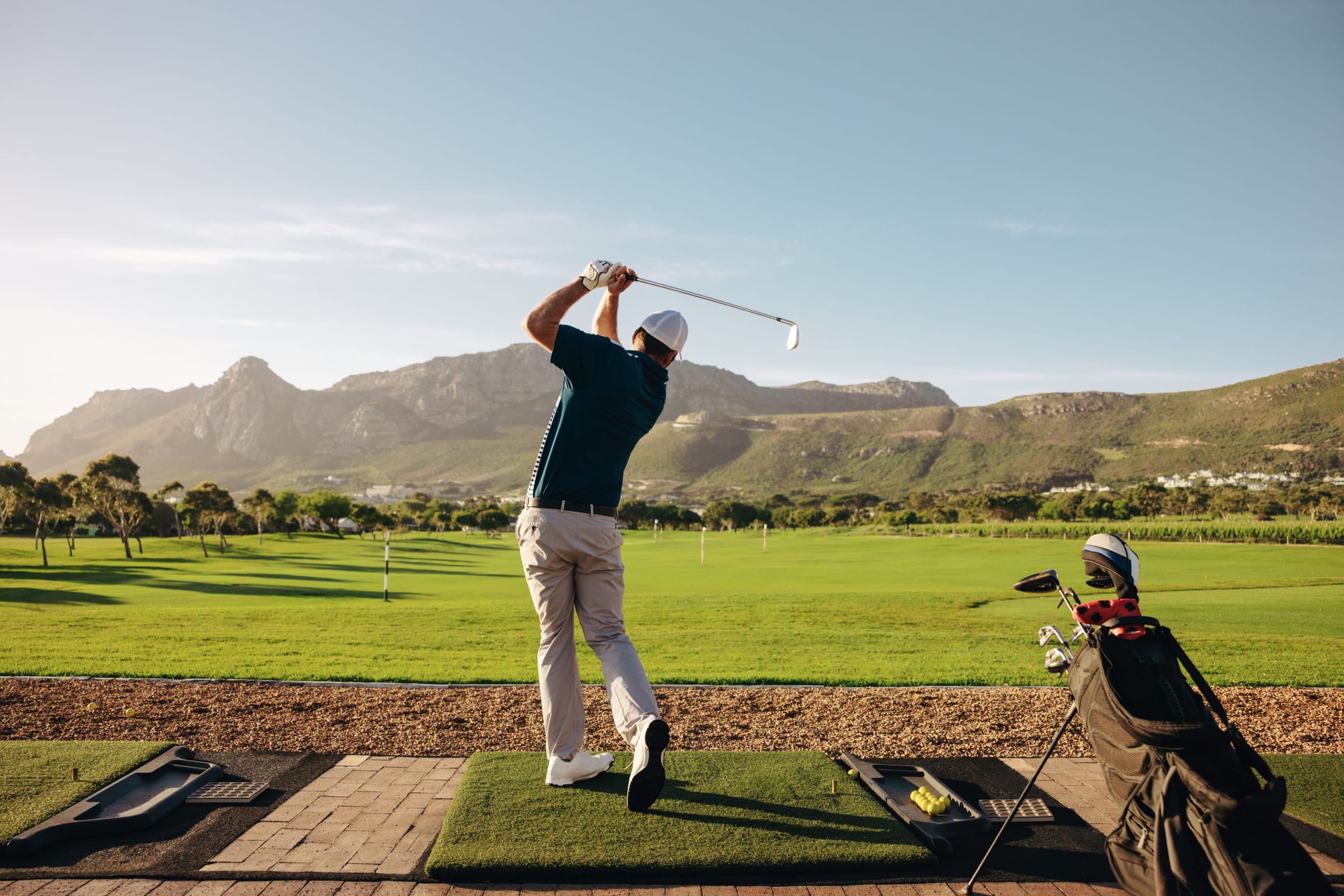 A golfer at the top of a full swing on a practice tee, with a mountain range beyond the fairway.