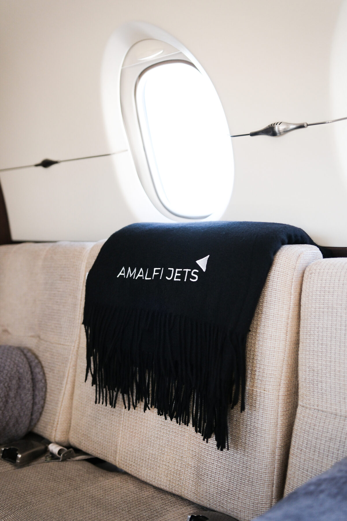 A black Amalfi Jets cashmere blanket draped over a cream cabin seat beside an oval jet window.