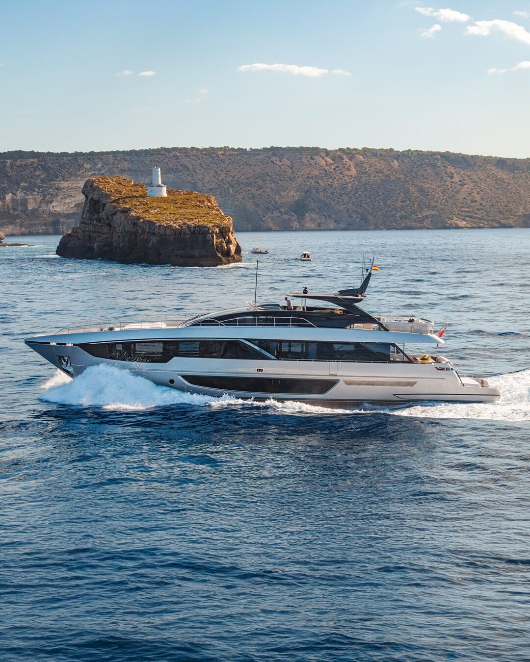 A superyacht under way past a rocky headland on open blue water.