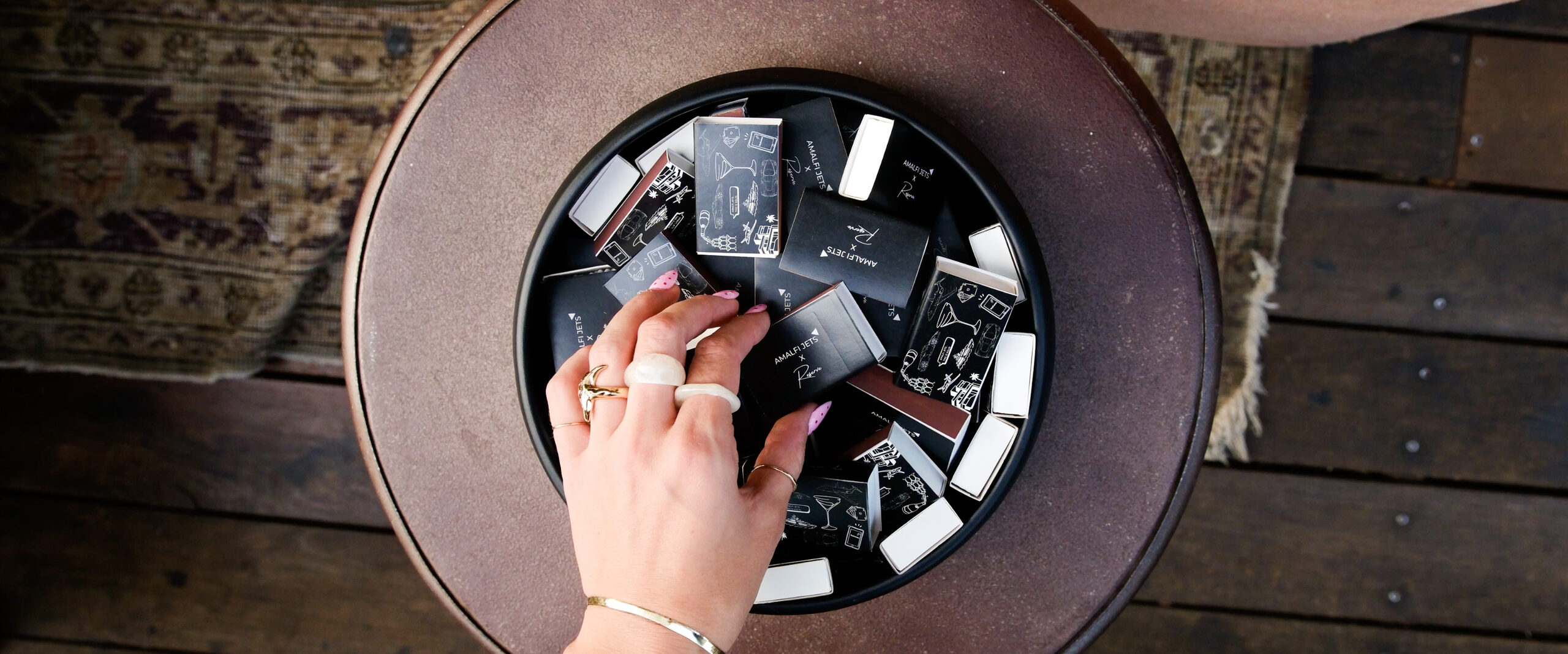 A hand reaching into a round leather case filled with Amalfi Jets matchboxes on a patterned rug.