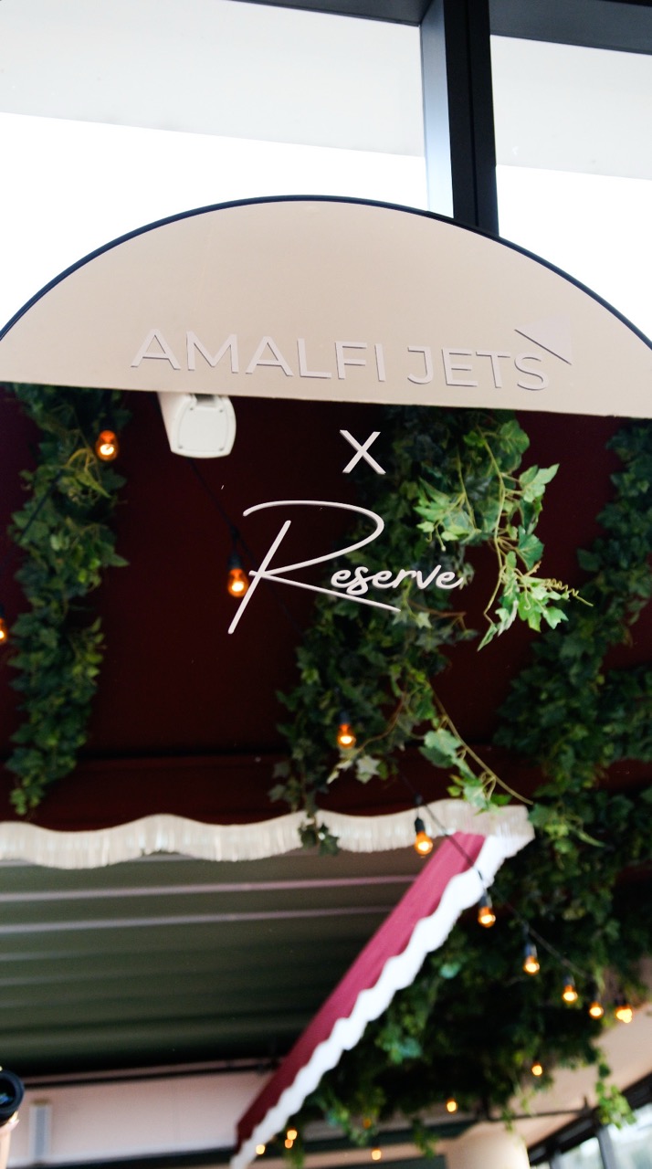 Tall standing mirror with Amalfi Jets and Amalfi Reserve logo with reflection of vines on a rooftop.