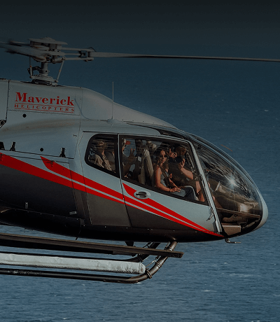 Black, gray, and red Maverick Helicopter flying over the ocean with passengers looking out the window.