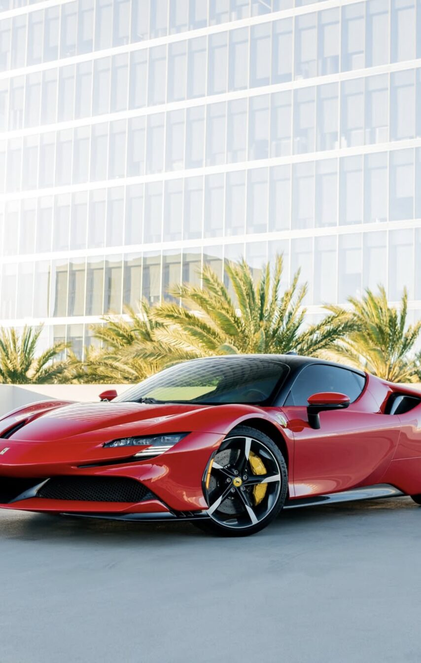 A red Ferrari parked beside a glass office tower lined with palm trees.
