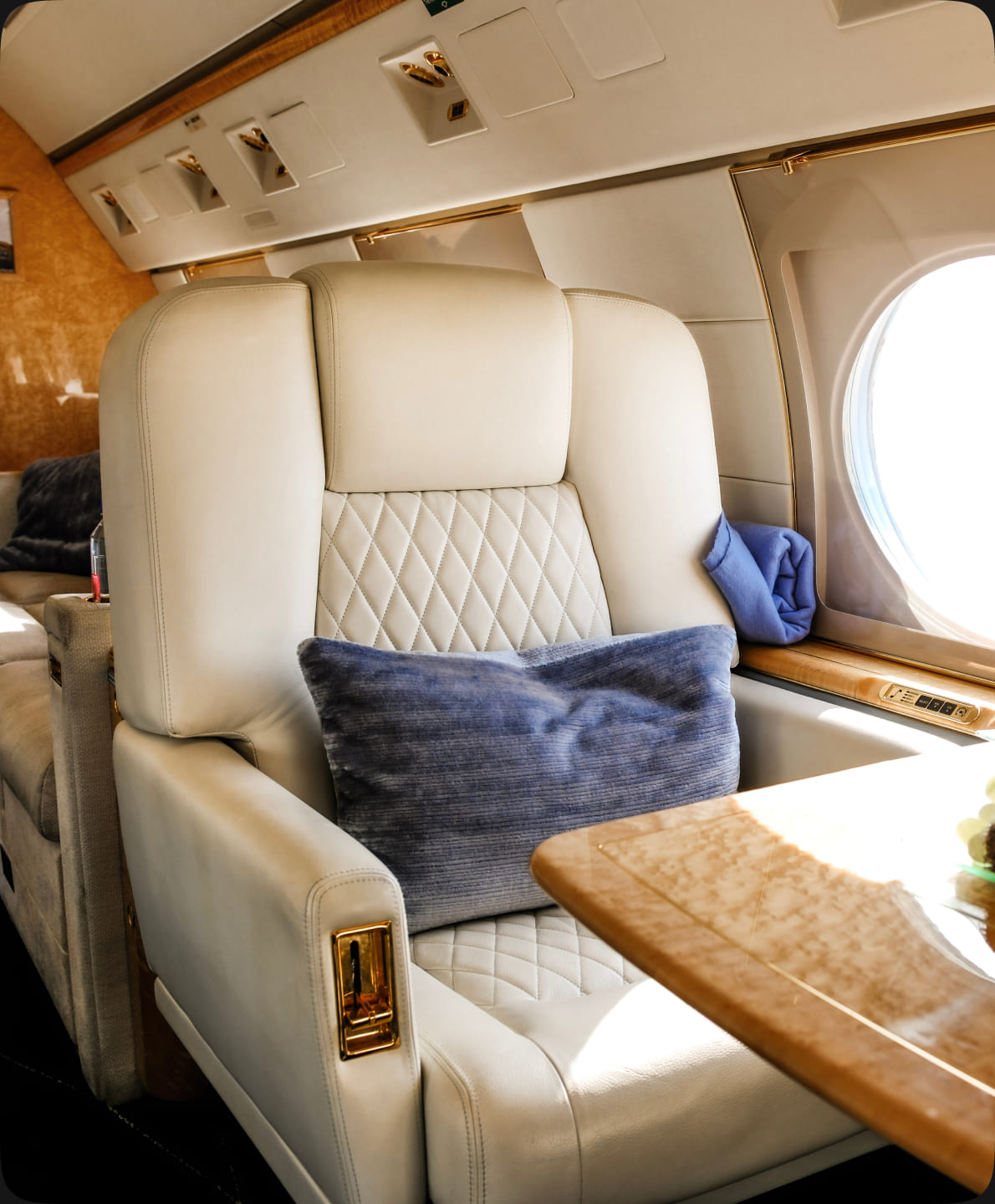 A cream leather seat with a blue cushion beside the wood table and oval window of a private jet cabin.
