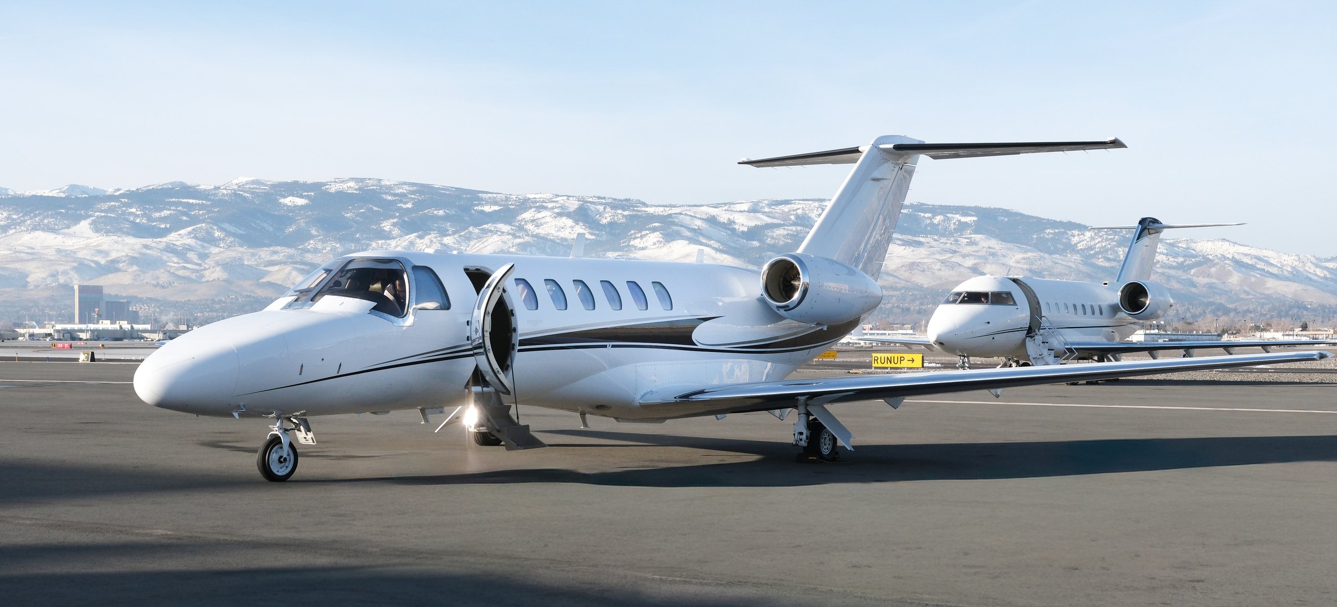 A private jet with its airstairs down on the tarmac, a second jet behind it and snow-covered mountains beyond.